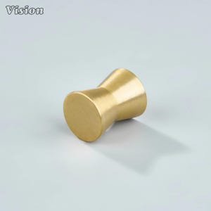 May include: A gold-colored cabinet knob with a unique hourglass shape. The knob has a smooth, matte finish and is designed with a cylindrical base and top, connected by a tapered center. The word "Vision" is in the top left corner.