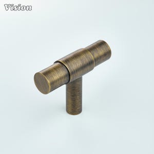 May include: Bronze-toned cabinet pull with a T-bar design. The hardware features a cylindrical horizontal bar connected to a vertical cylindrical post. The word "Vision" is printed in the upper left corner.