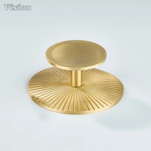 Puede incluir: Un pomo de armario dorado con una parte superior circular y una base más grande y texturizada. La base presenta un diseño de rayos de sol. La palabra "Vision" está en la esquina superior izquierda.