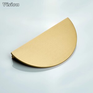 May include: A half-moon shaped cabinet pull in a brushed brass finish. The hardware has a flat, semi-circular design with a slightly curved edge. The word "Vision" is in the upper left corner.