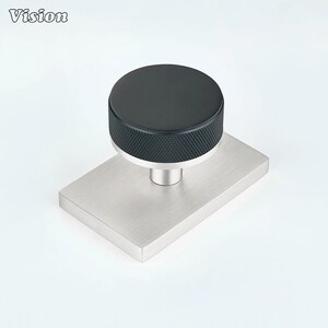 Pode incluir: Um puxador de móvel com topo circular preto com borda serrilhada texturizada e base de metal prateado. O puxador está montado numa placa retangular de metal prateado escovado. A palavra Vision está impressa no canto superior esquerdo.