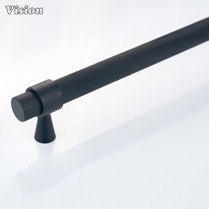 Peut inclure: Une poignée de meuble noir mat composée d'une longue barre cylindrique avec un motif texturé en croisillons. La poignée est soutenue par une base conique et un support de montage circulaire, offrant une apparence industrielle épurée.