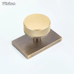 Op de afbeelding: Messingkleurige kastknop met een rechthoekige basis en een ronde, getextureerde bovenkant. De knop heeft een vintage uitstraling en is geschikt voor meubels of kasten. Het woord "Vision" staat in de linkerbovenhoek.