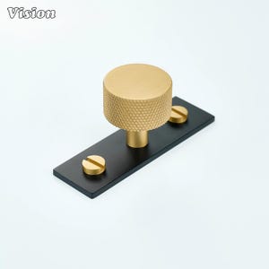 Op de afbeelding: Een goudkleurige kastknop met een getextureerde, cilindrische handgreep en een zwarte rechthoekige basis. Twee gouden schroeven bevestigen de knop aan de basis. Het woord "Vision" staat in de linkerbovenhoek.