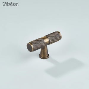 May include: A bronze-colored cabinet pull with a textured, cylindrical handle and a T-shaped design. The Vision logo is in the upper left corner. The hardware is made of metal and is suitable for drawers and cabinets.