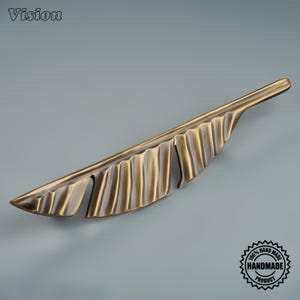 May include: A metallic cabinet handle shaped like a long, slender tropical leaf. The hardware features a brushed antique brass finish with dark, recessed grooves that highlight the rippled texture and organic, elongated form of the leaf design.