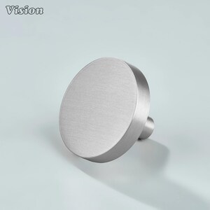 May include: A circular cabinet knob with a brushed silver finish. The knob features a flat, round face and a cylindrical base extending from the center of the back. The surface has a subtle metallic texture, reflecting light across its smooth, circular top.