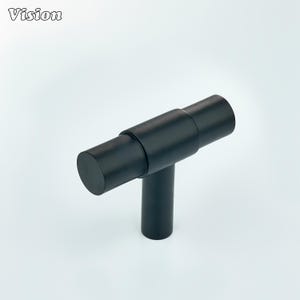 May include: A black T-bar cabinet handle. The cylindrical handle is connected to a vertical post. The word "Vision" is in the upper left corner. The handle is suitable for a variety of furniture styles.