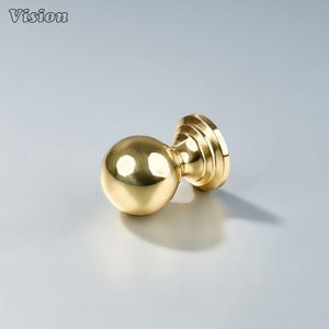 Puede incluir: Un pomo de puerta dorado brillante con un mango esférico y una base circular. El pomo de la puerta es de metal y tiene un acabado pulido. La palabra "Vision" está en la esquina superior izquierda.