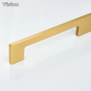 May include: A gold-colored cabinet pull with a rectangular design. The pull has a flat, bar-like shape with a right-angle bend at one end. The word "Vision" is in the upper left corner. This hardware is suitable for drawers and cabinets.