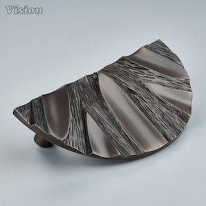 Puede incluir: Un tirador de mueble semicircular con acabado en bronce oscuro. La superficie presenta un patrón orgánico texturizado de crestas elevadas y ranuras hundidas que irradian hacia afuera, creando una apariencia dimensional en el tirador curvo.