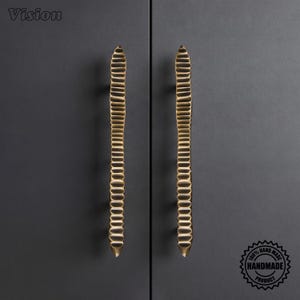 May include: Two long, vertical cabinet handles in a metallic gold finish. Each handle features a repeating, horizontal ribbed texture along its entire length, tapering to a point at both the top and bottom ends. The word Vision is in the top left.