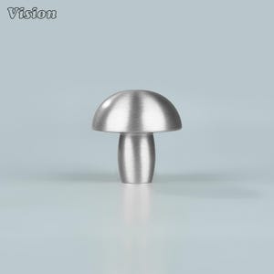以下が含まれることがあります： つや消し金属製のキノコ型キャビネットノブ。ノブは丸みを帯びた上部と円筒形のステムを備えています。「Vision」の文字が左上にあります。ノブはシルバー色で、水色の背景に配置されています。