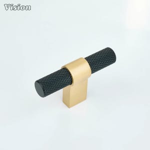 May include: A black and gold T-bar cabinet pull. The cylindrical handle is black with a textured pattern. The base and ring are gold. The word "Vision" is in the upper left corner.