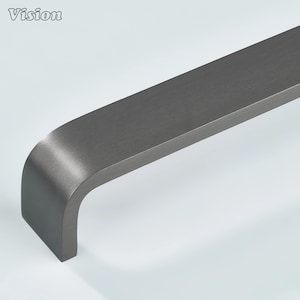 Op de afbeelding: Een donkergrijze metalen meubelgreep met een geborstelde afwerking en een gebogen uiteinde. Het woord Vision staat in de linkerbovenhoek in een wit lettertype met een zwarte omlijning. De greep heeft een lang, plat ontwerp met ronde randen.