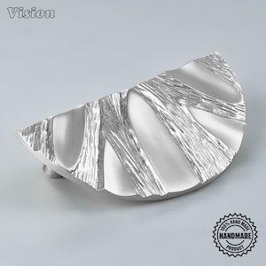 May include: A semi-circular silver cabinet pull with a textured finish featuring alternating smooth, polished sections and rough, striated patterns. The hardware has a curved, fan-like shape with a metallic sheen and a distinct, tactile surface design.