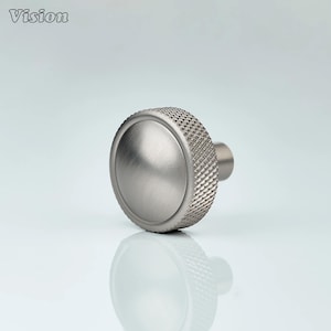 May include: A brushed metal cabinet knob with a textured, ridged edge and a smooth, circular face. The knob has a short, cylindrical stem. The word "Vision" is in the upper left corner.