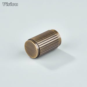May include: A cylindrical bronze-colored cabinet knob with a ridged center section and flat end caps. The word "Vision" is printed in the upper left corner. The knob is designed for furniture and home decor.