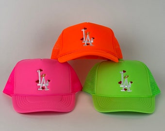 May include: Three brightly coloured trucker hats: neon orange, hot pink, and lime green. Each hat features the embroidered letters "LA" with a halo and hearts. The hats have a mesh back and a solid front.