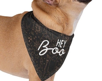 May include: A tan French Bulldog wearing a black bandana with a Halloween pattern. The bandana has the words "Hey Boo" in white script. The dog is looking up with its tongue out.