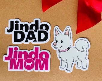 May include: Three stickers featuring a white dog with a pink collar, the text "Jindo DAD" in black, and the text "Jindo MOM" in pink with a small dog silhouette.