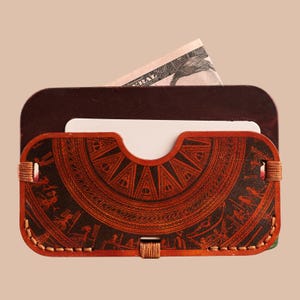 May include: A brown leather wallet with a decorative design and a stitched border. A white card and a portion of a U.S. dollar bill are visible inside. The wallet has a rectangular shape with rounded corners.