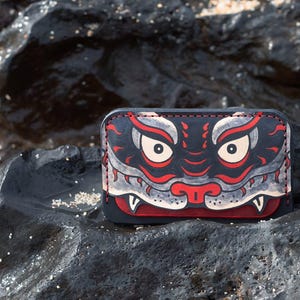 May include: A rectangular leather card holder featuring a stylised graphic of a creature face with large eyes, sharp teeth, and a nose in shades of black, red, and white. The item has visible stitching along the top edge and rounded corners.