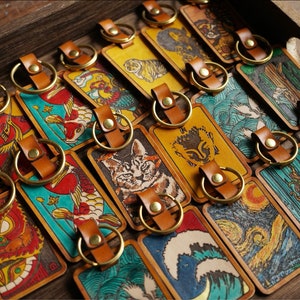 Peut inclure: Une collection de porte-clés en cuir avec différents motifs colorés, dont un chat, un tigre, une vague et un poisson-koi. Les porte-clés sont disposés dans un plateau en bois.