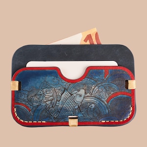 Puede incluir: Un tarjetero de cuero azul marino con un borde rojo y un diseño detallado de peces azules y blancos. El tarjetero tiene una ranura para tarjetas y un compartimento con un billete visible.