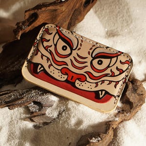 Może przedstawiać: Prostokątny skórzany wizytownik z ręcznie malowanym wzorem przedstawiającym stylizowaną twarz stworzenia z wyraźnymi oczami, czerwonymi i czarnymi akcentami oraz widocznymi przeszyciami wzdłuż krawędzi. Skóra ma jasnobeżowy kolor i gładkie wykończenie.