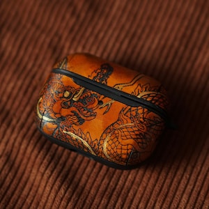 Könnte beinhalten: Ein braunes Leder-Airpods-Etui mit einem goldenen Drachen-Design.