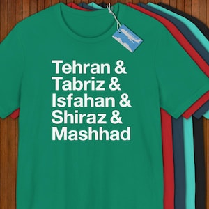 May include: A green t-shirt with the cities of Tehran, Tabriz, Isfahan, Shiraz, and Mashhad printed in white. The shirt has a crew neck and short sleeves. Several other t-shirts in different colors are visible behind the green shirt.