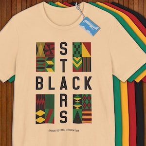May include: A cream-colored t-shirt with the text "STARS BLACK" in black block letters, with colorful geometric patterns. The shirt also has the text "GHANA FOOTBALL ASSOCIATION" in small letters. Several other t-shirts are stacked behind it.