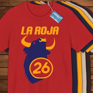 May include: Red t-shirt featuring the text "LA ROJA" in yellow, above a blue bull graphic with yellow horns and the number "26" in a yellow circle. Other t-shirts in various colours are visible.
