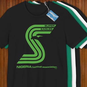 May include: Black t-shirt with a large green graphic of the words "SUPER EAGLES" and the letter "S" in a stylized design. The text "NIGERIA football association" is below the graphic. Several t-shirts are stacked.