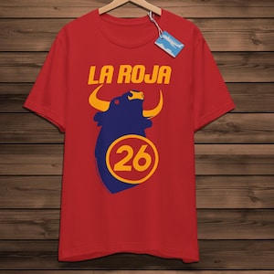 May include: Red t-shirt featuring the text "LA ROJA" in gold above a blue bull graphic with yellow horns. The number "26" is inside a yellow circle below the bull. The shirt is displayed on a wooden hanger.