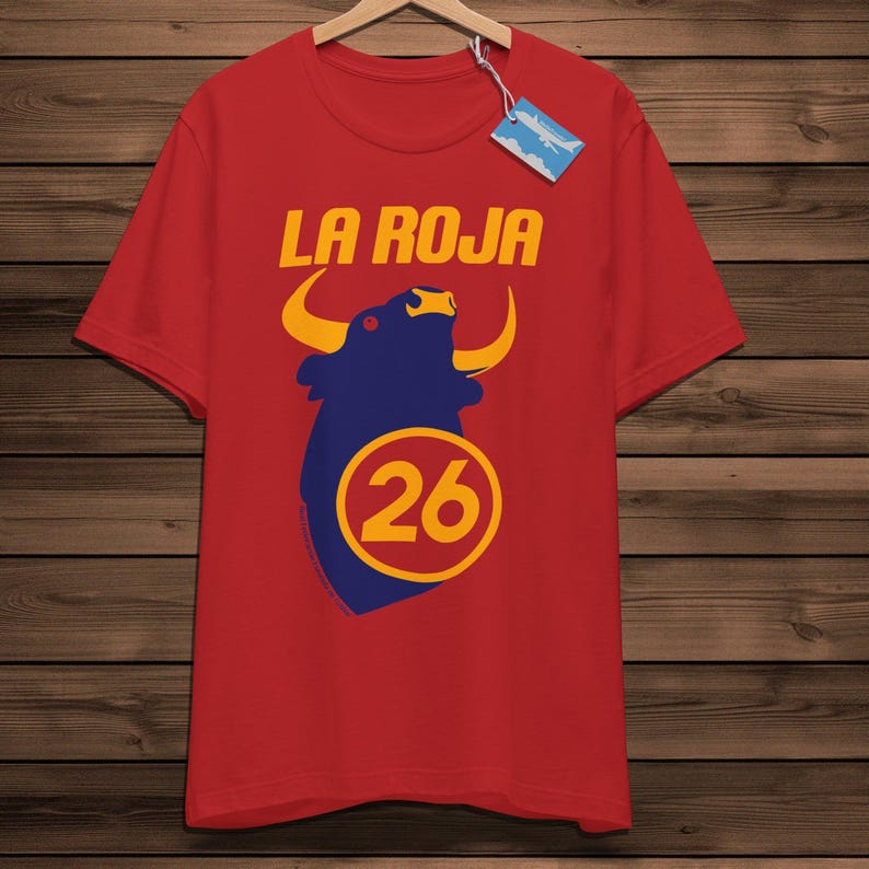 May include: Red t-shirt featuring the text "LA ROJA" in gold above a blue bull graphic with yellow horns. The number "26" is inside a yellow circle below the bull. The shirt is displayed on a wooden hanger.