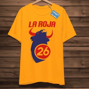 May include: Mustard yellow t-shirt featuring the red text "LA ROJA" above a navy blue bull graphic with red horns and the number 26 in a red circle. The shirt is displayed on a wooden hanger.