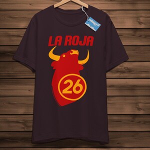 May include: A dark maroon t-shirt featuring the words "LA ROJA" in red above a red bull graphic with yellow horns. Below the bull is the number "26" in a yellow circle. The shirt is displayed on a wooden background.
