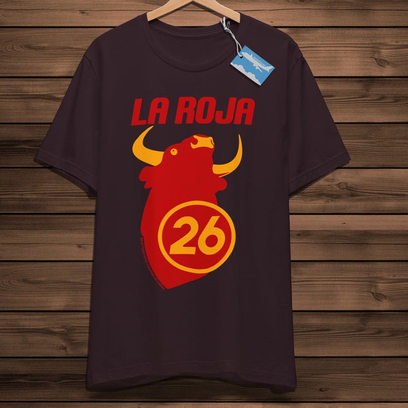 May include: A dark maroon t-shirt featuring the words "LA ROJA" in red above a red bull graphic with yellow horns. Below the bull is the number "26" in a yellow circle. The shirt is displayed on a wooden background.