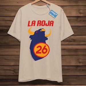 May include: A beige t-shirt featuring the text "LA ROJA" in red, above a blue bull graphic with yellow horns and a yellow and red circle with the number 26.