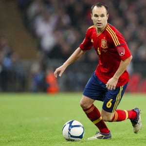 May include: A footballer in a red jersey with yellow stripes and the number 6 on the shorts and jersey. The player is wearing blue shorts, red socks, and white and black cleats. A football is at the player's feet.