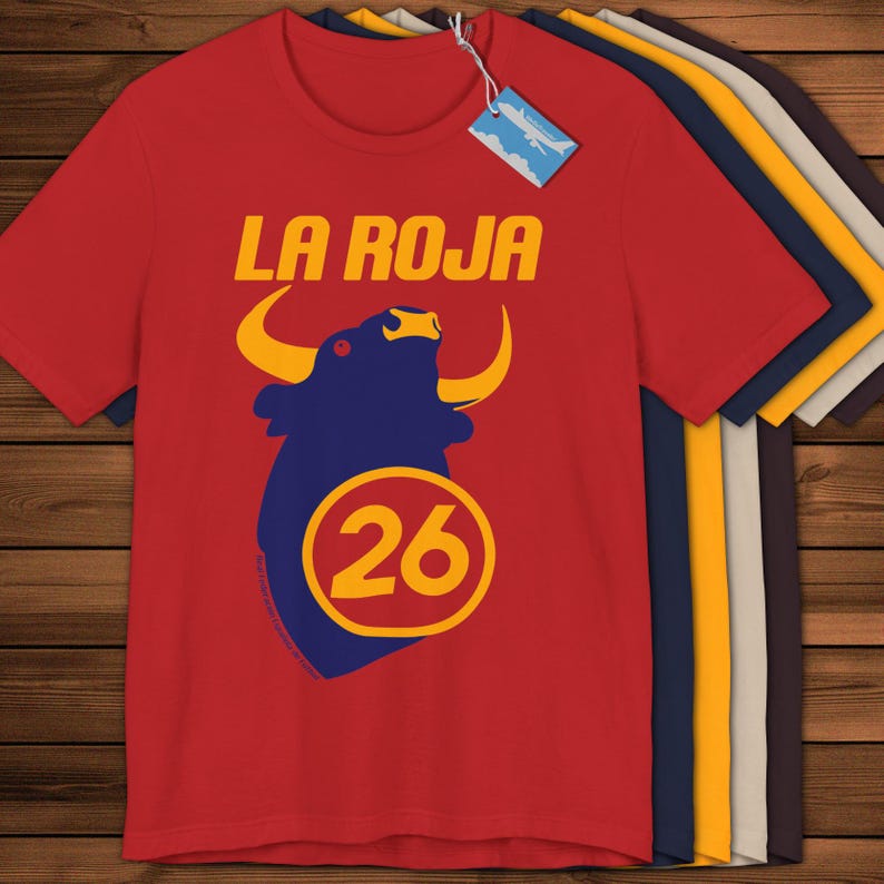 May include: Red t-shirt featuring the text "LA ROJA" in yellow, above a blue bull graphic with yellow horns and the number "26" in a yellow circle. Other t-shirts in various colours are visible.