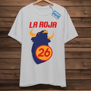 May include: A white t-shirt featuring the text "LA ROJA" in red. Below is a navy blue bull graphic with yellow horns and a yellow circle with the number 26. The shirt is displayed on a wooden hanger.