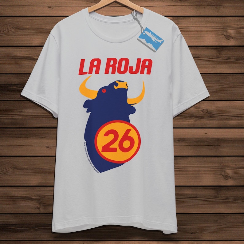 May include: A white t-shirt featuring the text "LA ROJA" in red. Below is a navy blue bull graphic with yellow horns and a yellow circle with the number 26. The shirt is displayed on a wooden hanger.