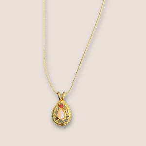 May include: A gold-toned necklace with a teardrop pendant. The pendant features a light pink gemstone and a small red accent stone, set within a filigree design. The chain is delicate and the overall style is vintage.