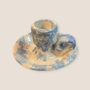 Könnte beinhalten: Handgefertigter Kerzenhalter aus Keramik mit blau-weißem Sprenkelmuster. Der Kerzenhalter hat einen kreisförmigen Sockel, eine zentrale Tasse für die Kerze und einen kleinen Griff. Die Farbpalette ist eine Mischung aus Hellblau und Creme.