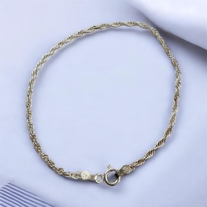 Puede incluir: Una pulsera de cadena de cuerda de color plateado con un cierre de mosquetón. La pulsera está hecha de hebras de metal retorcidas, creando una apariencia texturizada. El cierre está unido a la cadena con un pequeño anillo. La pulsera se muestra sobre una superficie blanca.