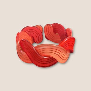 May include: A vibrant, abstract bracelet in shades of red and orange. The bracelet features a series of interconnected, curved segments with a striped pattern, creating a flowing, artistic design. The bracelet is a statement piece.