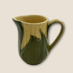 May include: A ceramic jug with a corn-on-the-cob design. The jug has a yellow interior, a handle, and a green exterior with a textured corn pattern. The jug is approximately 15 cm tall.